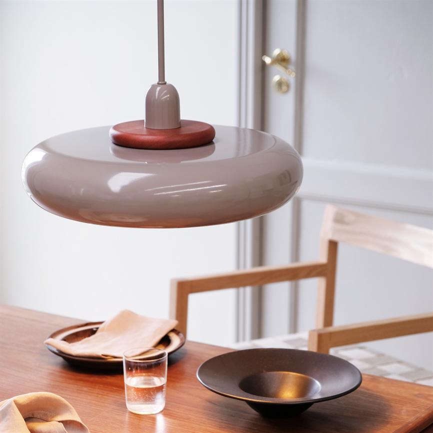 En rund, beige pendel hænger over et træspisebord; bordet rummer en sort skål, en tallerken med serviet, et glas vand og en træstol foran en hvid dør.