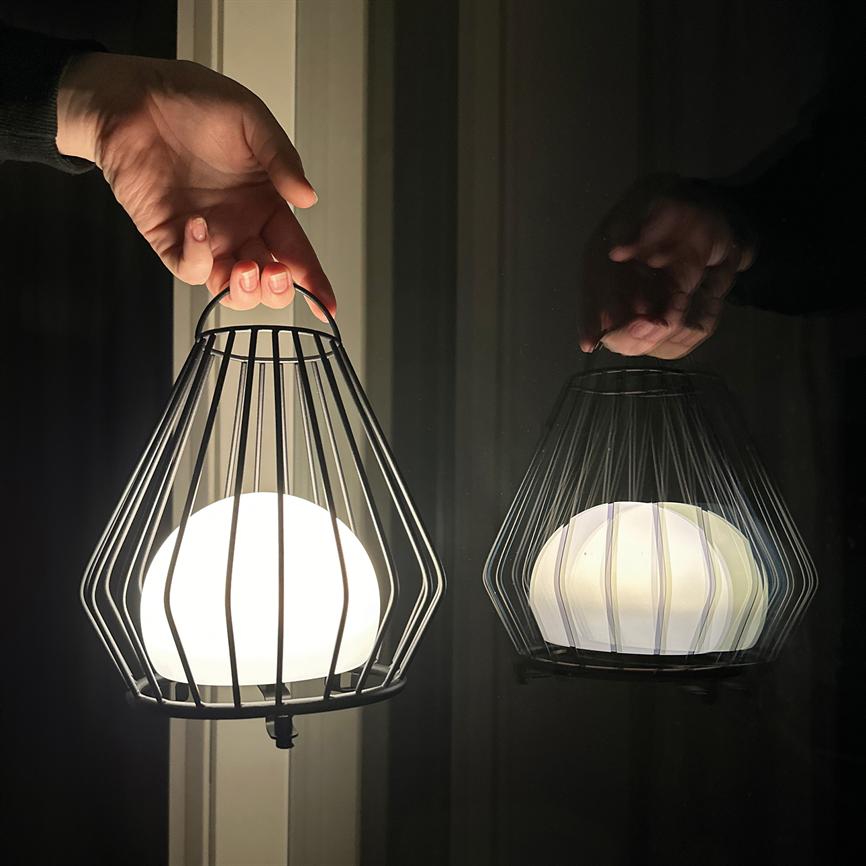Eine kugelförmige Lampe, die von einem Metallgitter umgeben ist, wird von einem Griff gehalten; Es strahlt warmes Licht aus und sowohl Lampe als auch Hand spiegeln sich in einer dunklen Fensterscheibe.