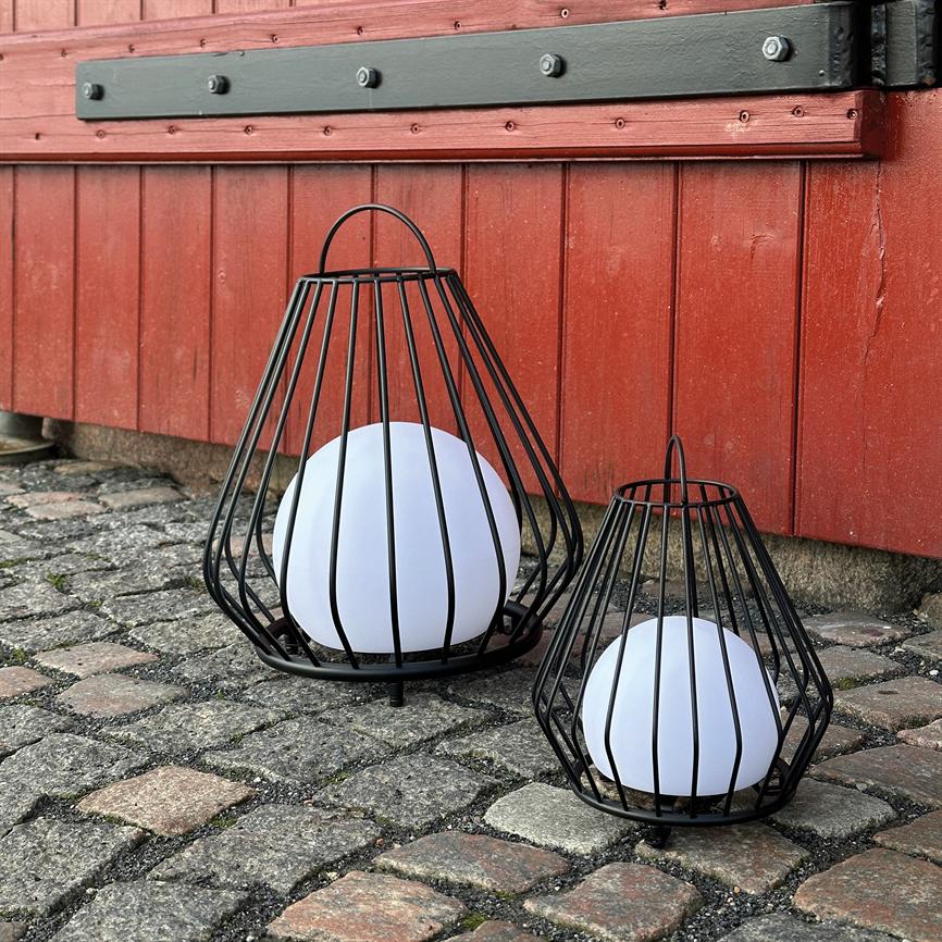 Zwei schwarze Metalllaternen, eine große und eine kleinere, halten weiße Kugellampen vor einem roten Holzgebäude, das auf einer gepflasterten Fläche steht.