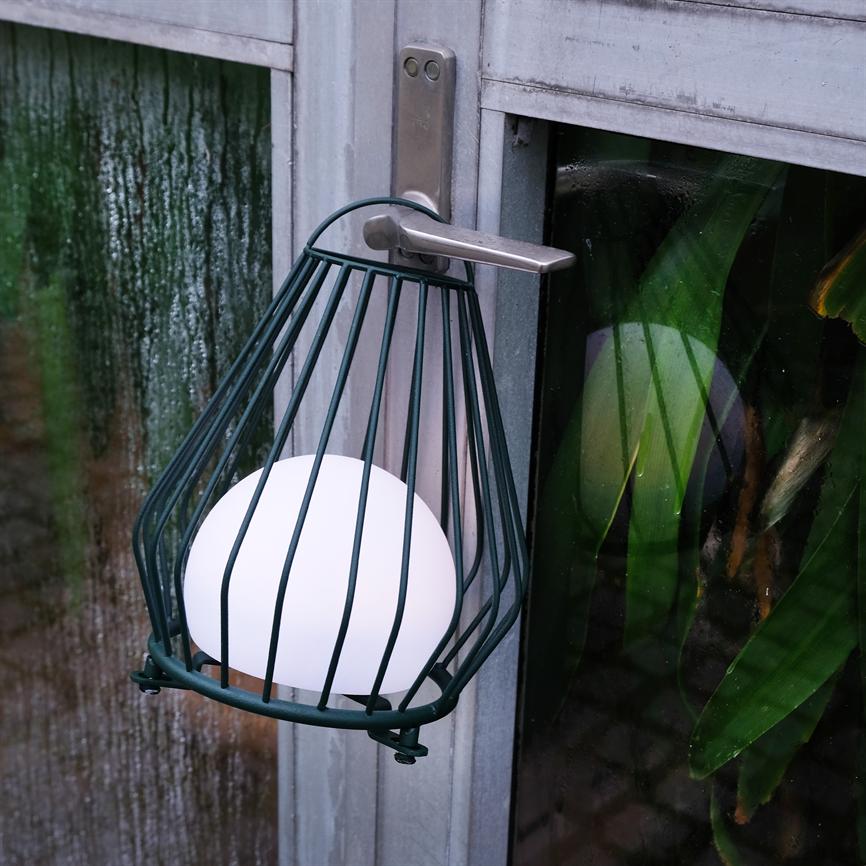 En lampe i metalbur med en hvid kuglelyspære hænger på et metal dørhåndtag; bagved dugget glas med refleksioner og grønne planter.