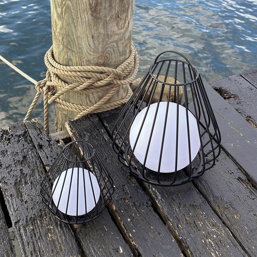 Zwei schwarze Metalllampen mit weißen runden Einsätzen ruhen auf einem abgenutzten, dunklen Holzdeck an einer an einem Seil befestigten Stange, im Hintergrund ruhiges Meerwasser.
