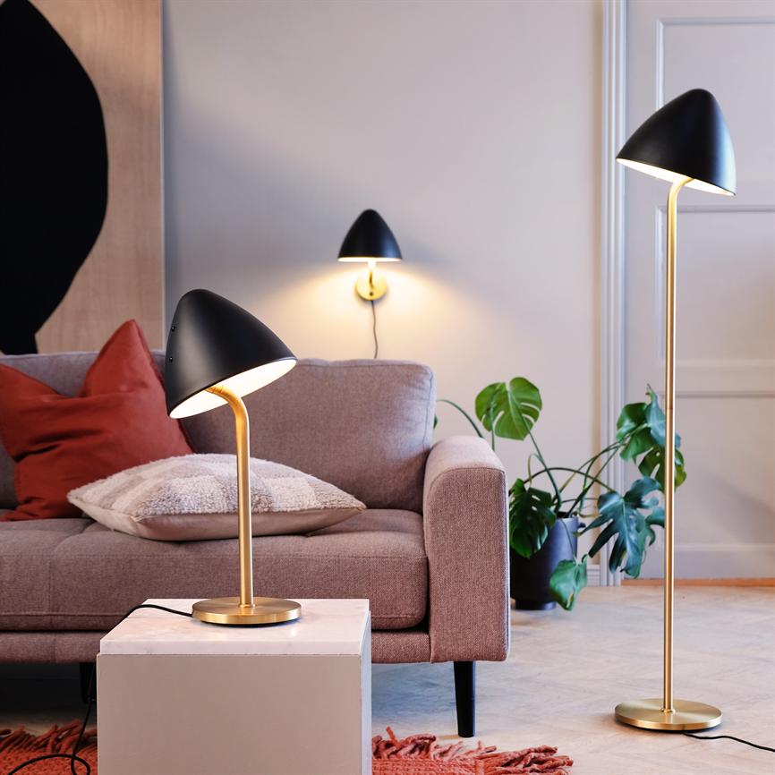 Neben einem gepolsterten Sofa leuchtet eine schwarz-goldene Tischlampe; ergänzt durch eine passende Stehleuchte und Wandleuchte, umgeben von Pflanzen, Kissen und einem warmen, gemütlichen Wohnraum.
