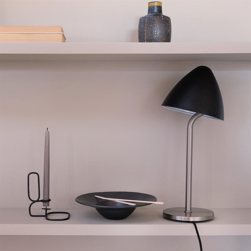 Rechts steht eine schwarze Tischlampe auf einem hellen Regal neben einer Schale mit Stöcken und einem Kerzenständer; Oben an einer neutralen Wand stehen eine Vase und Bücher.