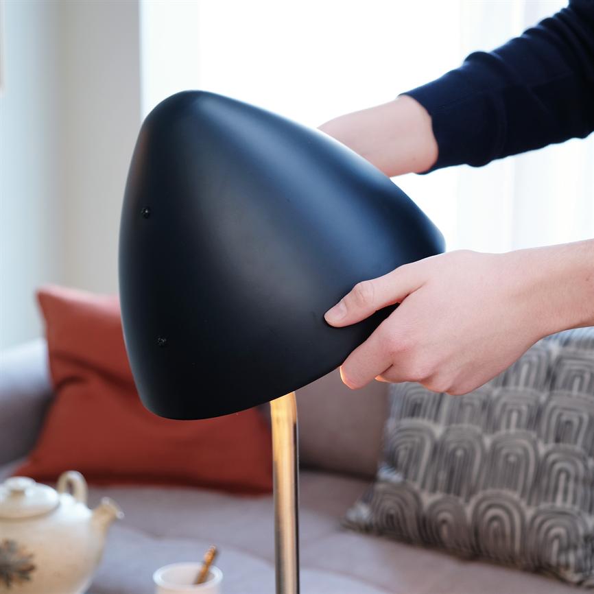 Sort bordlampe med rund skærm, justeres af to hænder; i en lys stue foran en sofa med rød og mønstret pude samt en tekande på bordet.