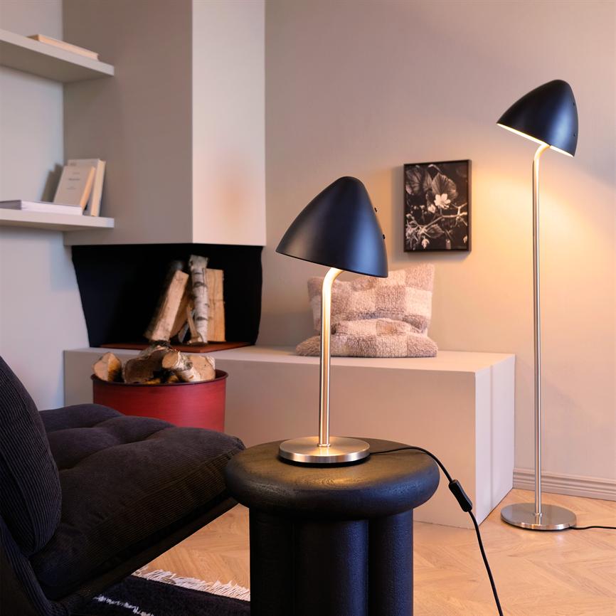 En sort bordlampe står tændt på et rundt sort sidebord og kaster varmt lys; i konteksten en hyggelig hjørnestue med matchende gulvlampe, mørk sofa, brændekurv og pude på lav hylde.