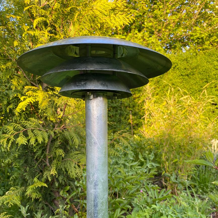Eine runde, gestufte, oben montierte Gartenlampe steht auf einem schlanken Metallmast zwischen sonnenbeschienenen Büschen und grünen Pflanzen in einem üppigen Garten.
