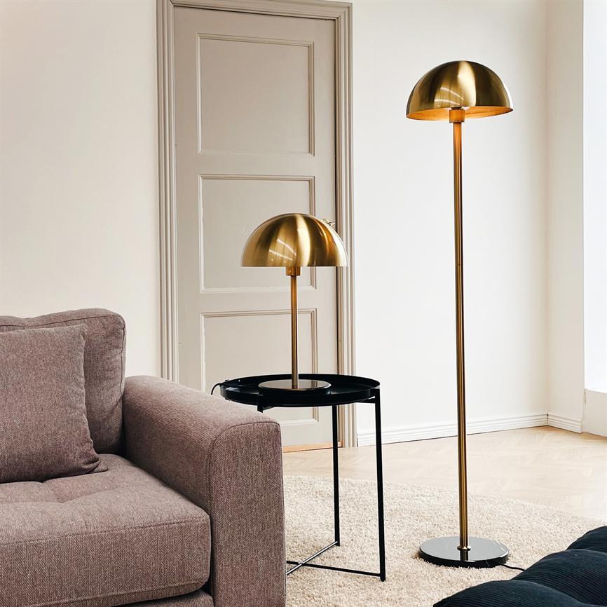 Zwei goldene Lampen stehen neben einem grauen Sofa; eine auf einem schwarzen Beistelltisch, die andere als Stehlampe auf dem Holzboden, umgeben von hellen Wänden, einer beigen Tür und einem hellen Teppich.