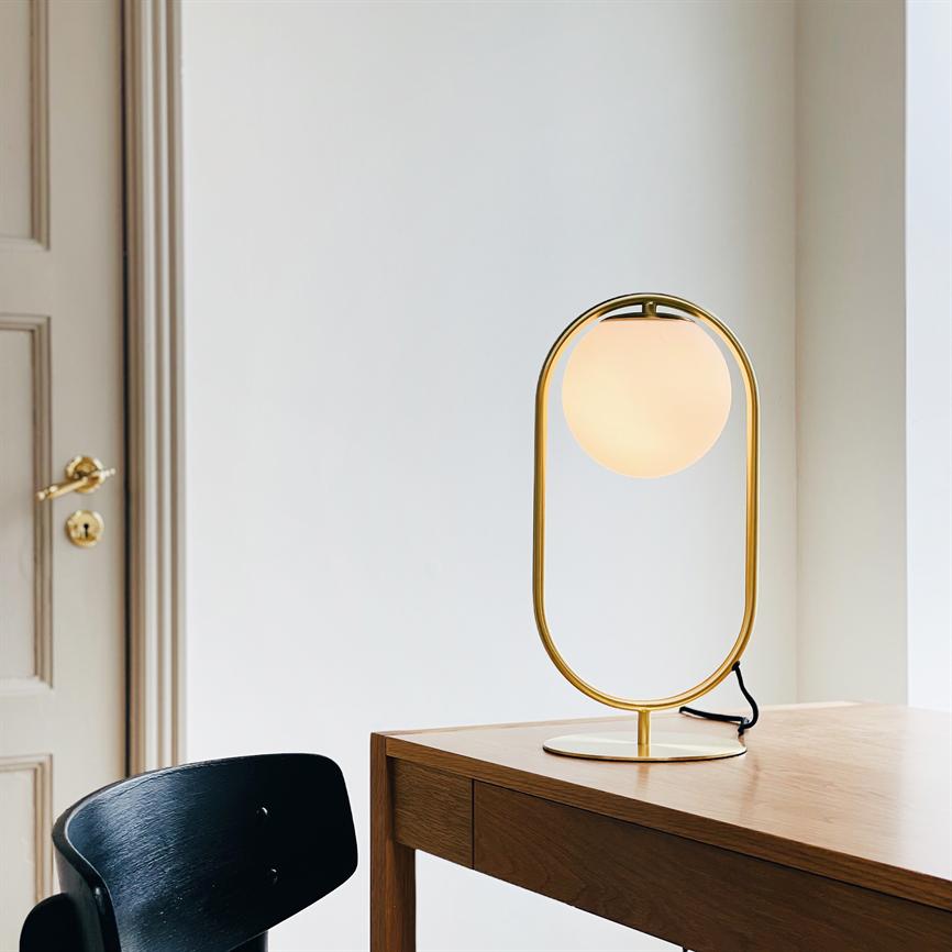 Eine goldfarbene Tischlampe mit ovalem Rahmen und rundem Glühlampenschirm; Es wird auf einem Holztisch von einem schwarzen Stuhl vor einer weißen Wand und einer Tür mit einem Messinggriff beleuchtet.