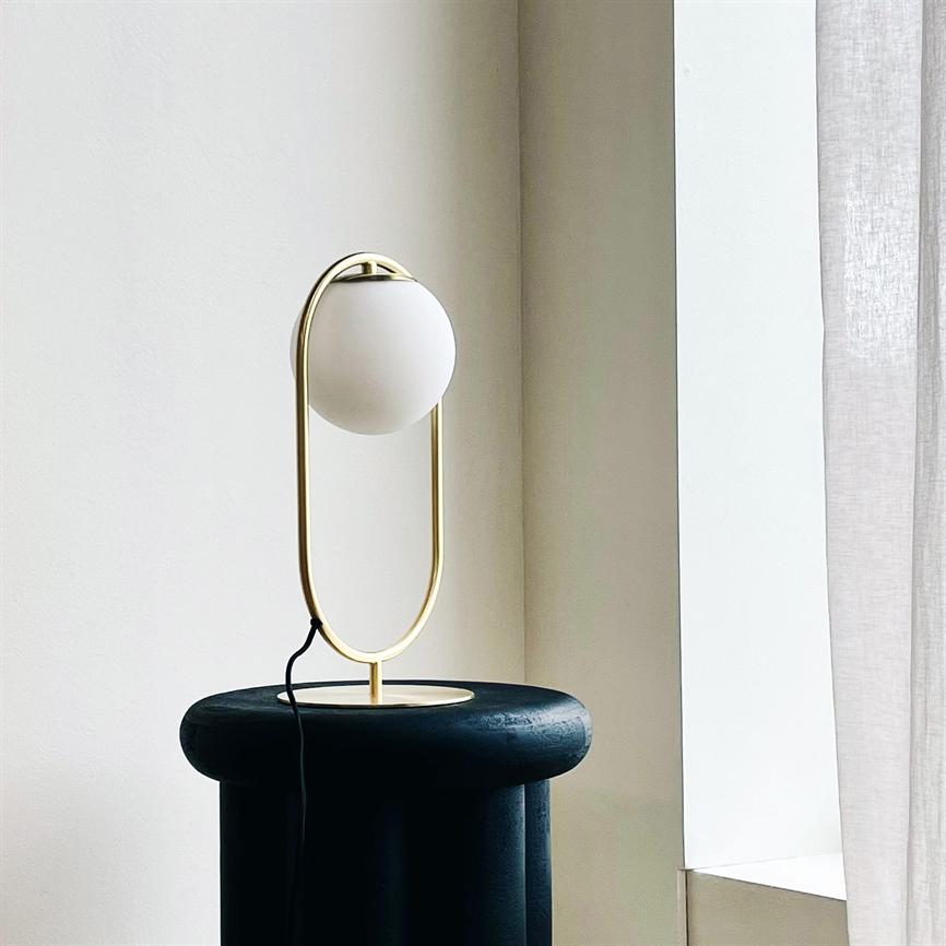 Eine moderne Tischlampe mit einer weißen Glaskugel in einem vergoldeten ovalen Rahmen wird auf einem schwarzen runden Hocker durch eine helle Fensterecke mit beigen Wänden und einem weißen Vorhang ausgeschaltet.