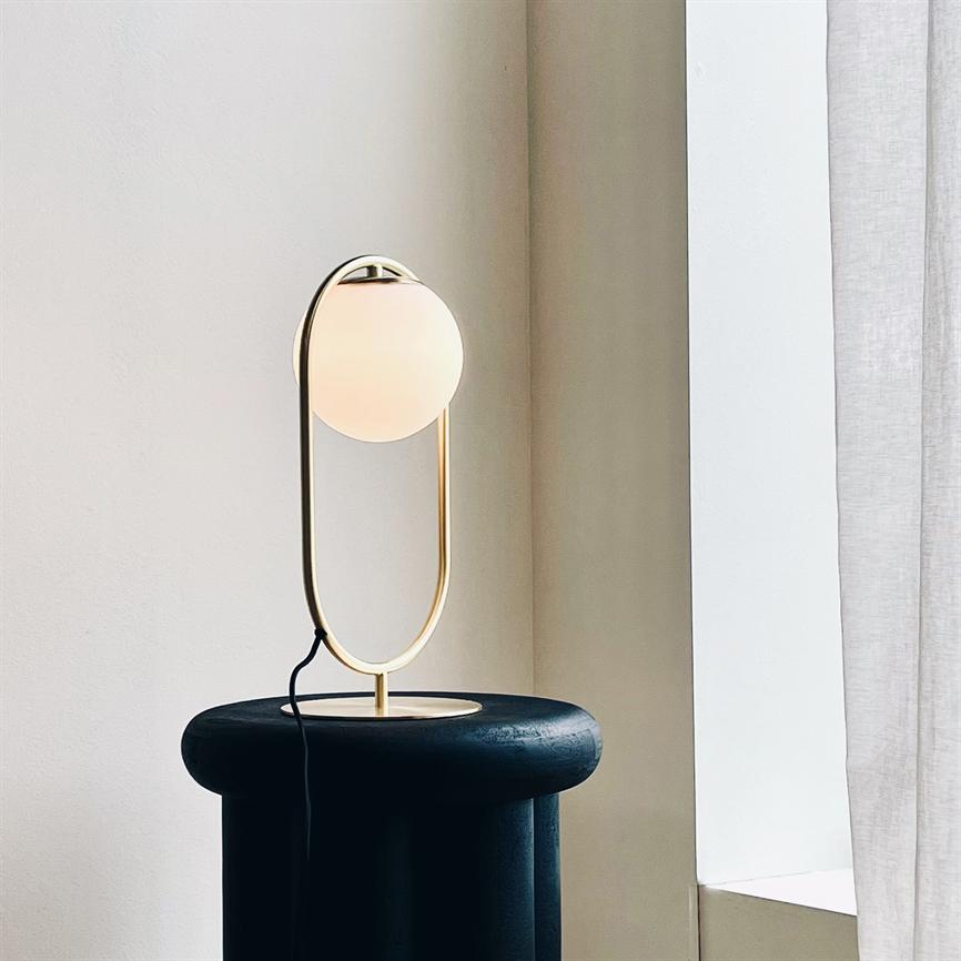 Eine moderne Tischlampe mit goldenem ovalem Fuß und Opalglaskugel strahlt weiches, warmes Licht aus und steht auf einem runden, dunklen Hocker in einer hellen Ecke an einem vorgehängten Fenster.