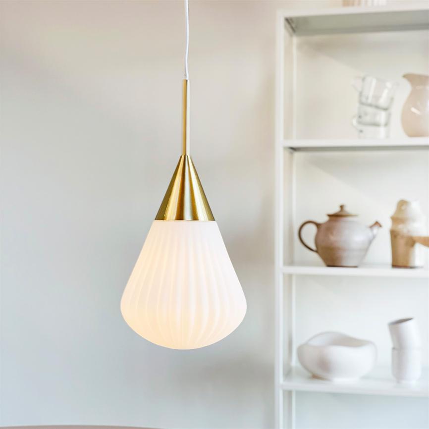 Ein weißes, zerrissenes pendel mit goldfarbener Oberseite hängt und spendet sanftes Licht vor einem weißen Bücherregal mit Keramikkrügen, -schalen und -bechern.