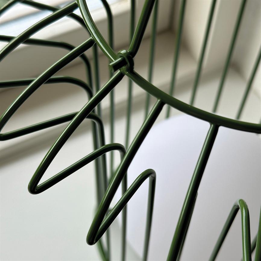 Der grüne Metalldrahtkorb bildet blattartige, geschwungene Fäden und steht neben einer Fensterbank in sanftem Tageslicht mit neutralem, verschwommenem Hintergrund.