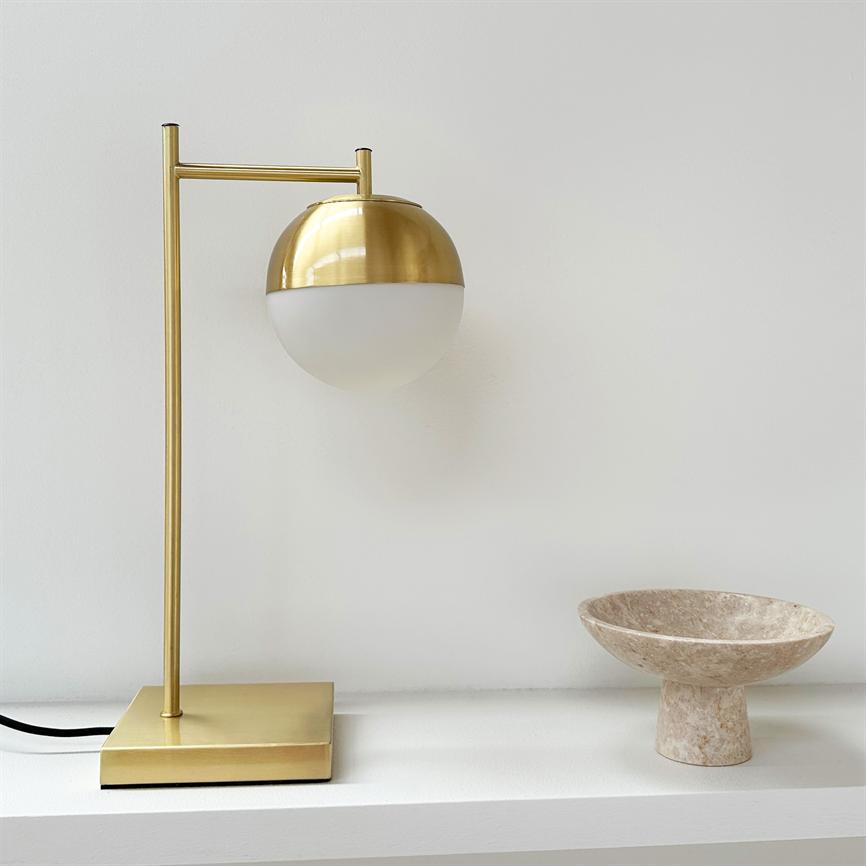 Goldfarbene Tischleuchte mit halbkugelförmigem Opalglas ist auf einem schlanken Stab montiert und steht auf einem quadratischen Sockel auf einem weißen Regal; rechts eine beige Marmorschale.