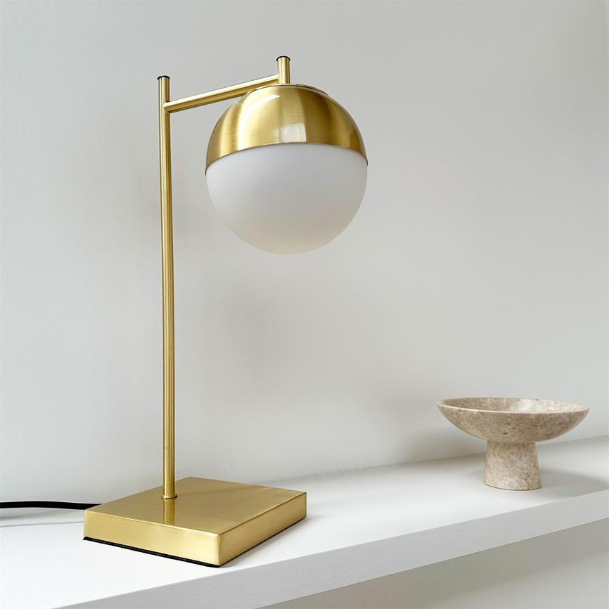 Goldene Tischlampe mit halbkugelförmigem weißen Schirm hängt an einem schlanken Stab über einem quadratischen Messingsockel; auf einem weißen Regal vor der Wand platziert, neben einer beigen Marmorschale.