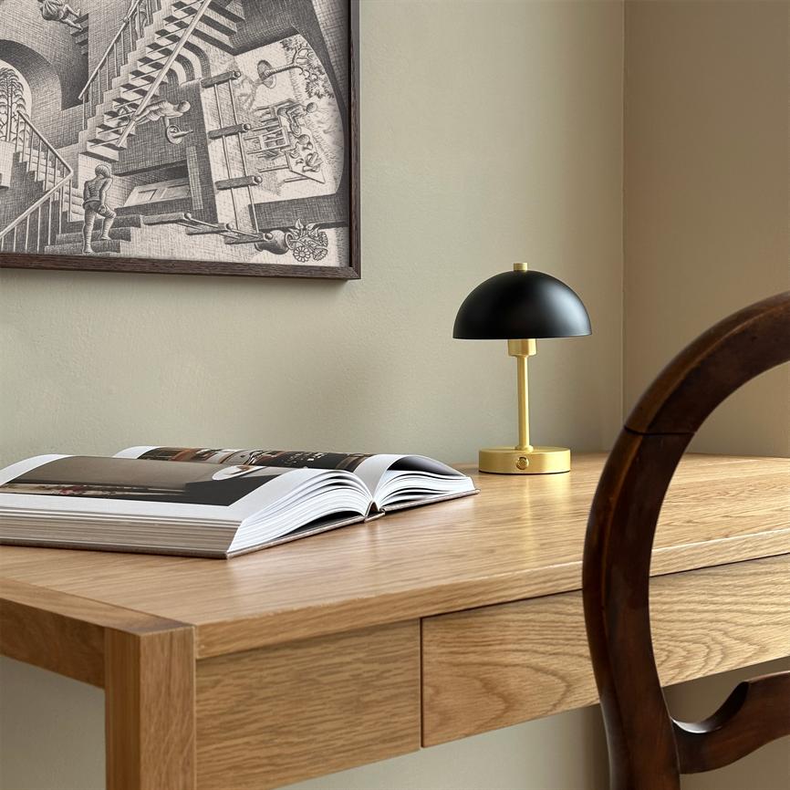 Auf einem hellen Holzschreibtisch liegt ein aufgeschlagenes Buch; Daneben ist eine kleine schwarz-goldene Tischlampe ausgeschaltet, an der Wand hängt ein grafisches Schwarz-Weiß-Poster und ein Holzstuhl wirft Schatten.