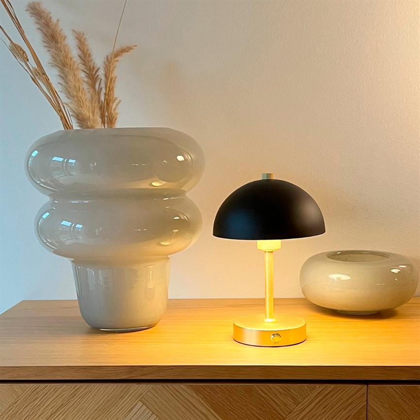 Großer glänzender Keramiktopf mit getrocknetem Pampasgras, eine kleine schwarz- und messingfarbene Tischlampe, die warm leuchtet, und eine niedrige runde Vase auf einem Holzschrank vor einer neutralen Wand.