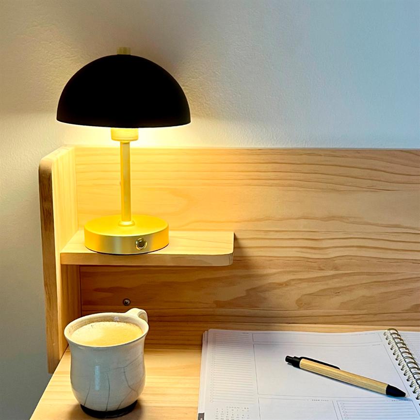 Eine gelbe Tischlampe mit schwarzer Kuppel leuchtet warm über einem hellen Holzschreibtisch; Links steht eine Keramiktasse mit Kaffee, rechts ein aufgeschlagenes Spiralnotizbuch mit Stift.