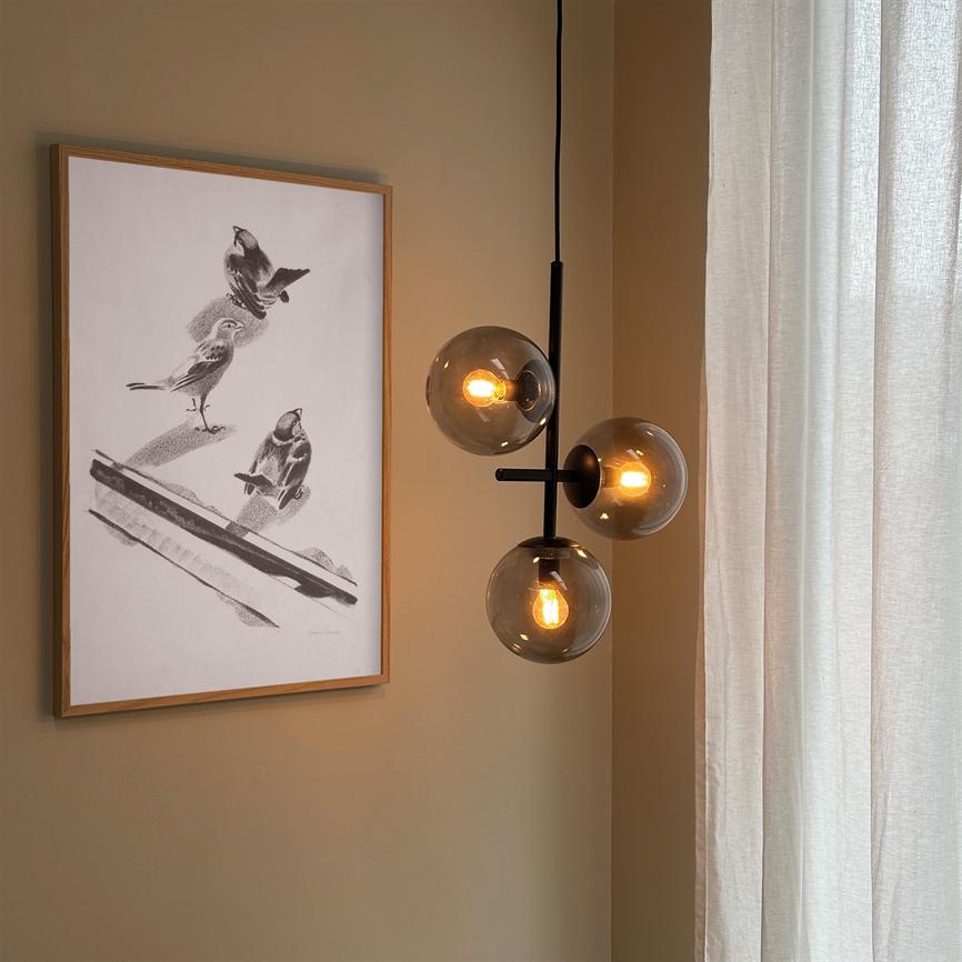 Eins pendel mit drei rauchigen Glaskuppeln leuchtet und strahlt warmes Licht vor einer beigen Wand mit einer gerahmten schwarz-weißen Vogelzeichnung und einem hellen Vorhang aus.