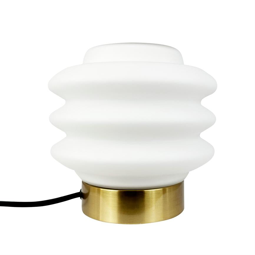 Eine weiße, gewellte Tischlampe ruht auf einem runden, gebürsteten Messingsockel; Das schwarze Stromkabel läuft seitlich vor einem neutralweißen Hintergrund heraus.