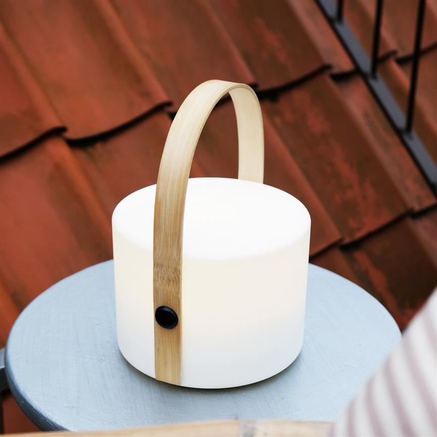 Weiße zylindrische Lampe mit gebogenem Holzgriff steht auf einem kleinen blauen runden Tisch. Sie strahlt sanftes Licht aus und wird vor roten Dachziegeln in einer Außenbalkonumgebung platziert.