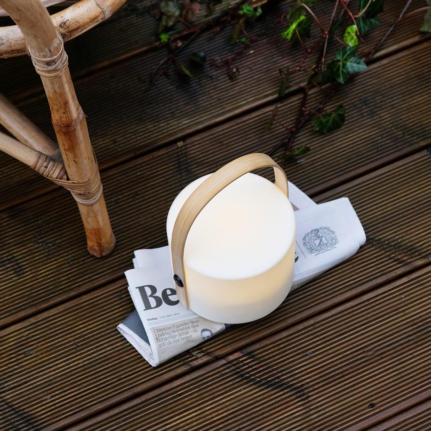 Runde tragbare Lampe mit Bambusgriff strahlt sanftes Licht aus, steht auf einer gefalteten Zeitung auf einer dunklen Holzterrasse neben einem Rattanstuhl und kletterndem Efeu. Text: „Bete“