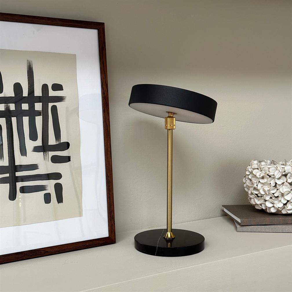 Eine kleine Bordlampe mit einem runden Rand und einer starken Messingstruktur, die sich auf eine neutrale Farbe stützt, von einem abstrakten Hintergrund, einem Baum und einer großen Skulptur flankiert wird.