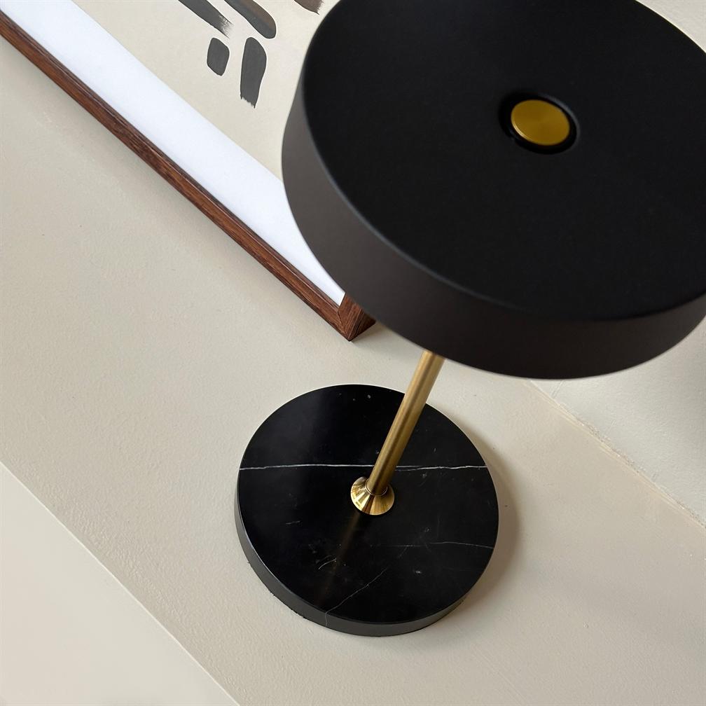 Black table lamp stands on a beige table; ein goldener Metallstab verbindet einen runden schwarzen Schirm mit einem runden schwarzen Marmorsockel; Dahinter hängt ein gerahmtes abstraktes Poster mit schwarzen Pinselstrichen.