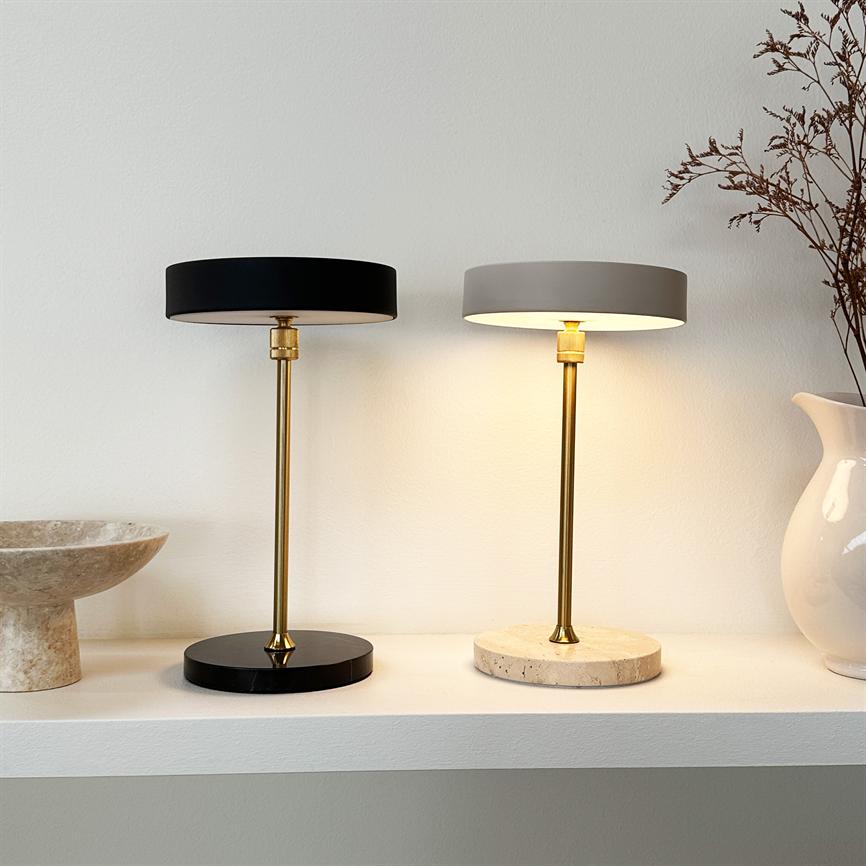 Zwei Tischlampen stehen nebeneinander: rechts an, links aus; beide mit Messinggriffen und runden Sockeln, auf einem weißen Regal an einer hellen Wand zwischen Schüssel und Vase platziert.