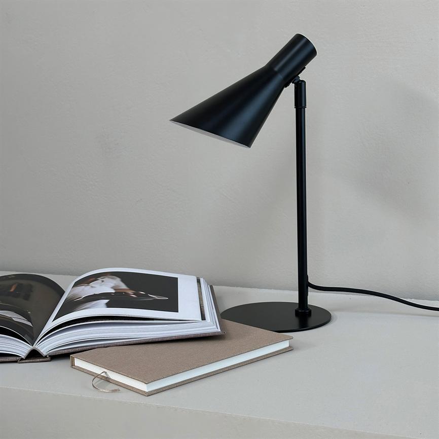 Eine Tischlampe aus schwarzem Metall zeigt nach unten über ein aufgeschlagenes Fotobuch; daneben liegt ein geschlossenes Notizbuch auf einer hellen Tischplatte mit sichtbarer Kordel vor einer hellgrauen Wand.
