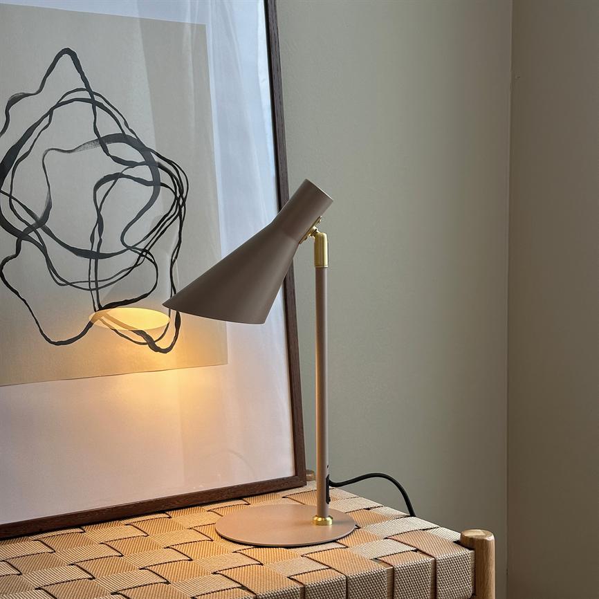 Eine Tischlampe in mattem Beige wirft warmes, goldenes Licht auf eine gerahmte abstrakte Strichzeichnung und eine Korbmatte vor einer hellen, neutralen Wand.