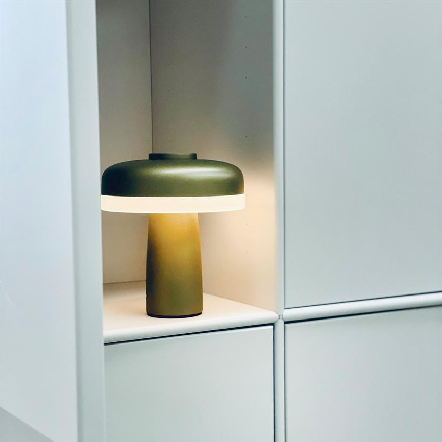 Auf einem Regal in einer weißen Schranknische brennt eine Tischlampe; Olivgrüner runder Schirm und zylindrischer Sockel, weiches, warmes Licht wirft Schatten auf die Rückwand.