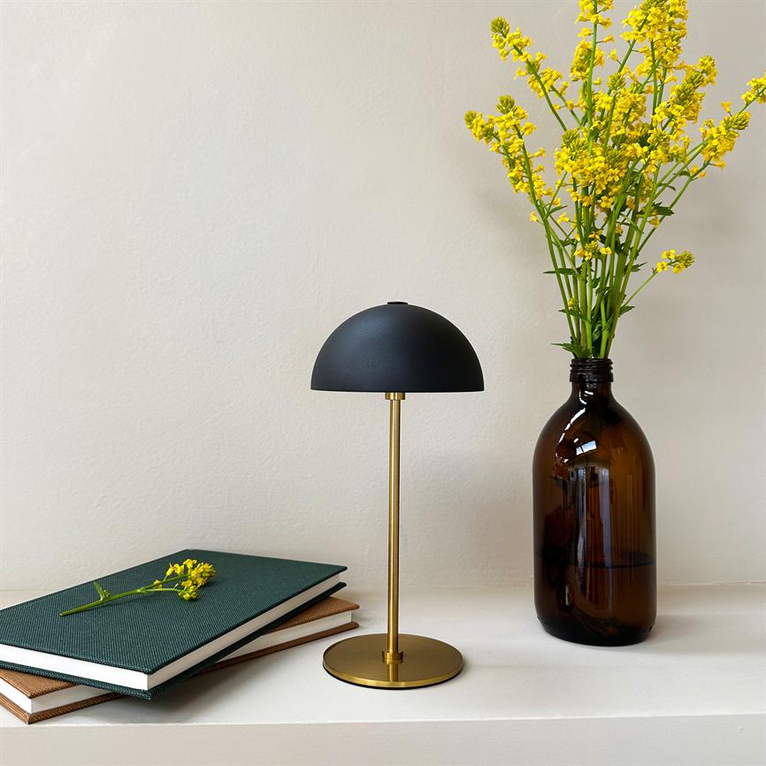 Eine kleine Tischlampe mit schwarzer Kuppel und goldenem Sockel steht auf einem weißen Regal zwischen zwei Büchern und einer braunen Glasflasche mit gelben Blumen vor einer hellen Wand.