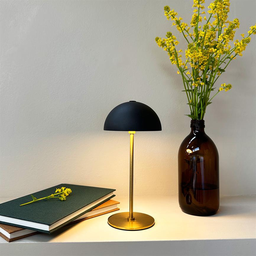 Lampe mit schwarzer Kuppel auf Messingsockel strahlt warmes Licht auf einem weißen Regal aus; daneben ein Stapel Notizbücher mit einer Blume und eine braune Flaschenvase mit gelben Blumen.