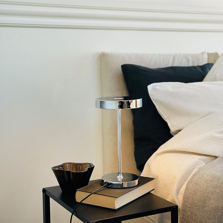 Tischlampe steht auf einem Buch auf einem schwarzen Nachttisch; daneben eine dunkle Schüssel; Der Nachttisch steht neben einem beigen Bett mit heller, gestreifter Bettwäsche und einem schwarzen Kissen.