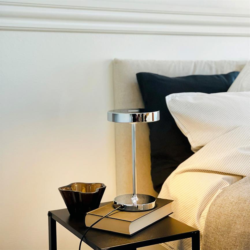 Tischlampe steht auf einem Buch auf einem kleinen schwarzen Nachttisch; Das Chrom der Lampe reflektiert das Licht. Daneben eine braune Keramiktasse, ein beiges Kopfteil, gestreifte weiße Bettwäsche und ein schwarzes Kissen im Hintergrund.