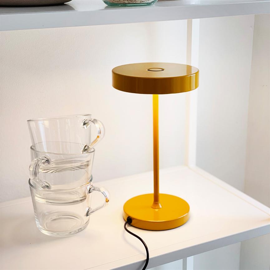 Eine gelbe Tischlampe leuchtet sanft auf einem weißen Regal; daneben stehen drei gestapelte Glasbecher, und vom Sockel der Lampe verläuft eine schwarze Schnur.