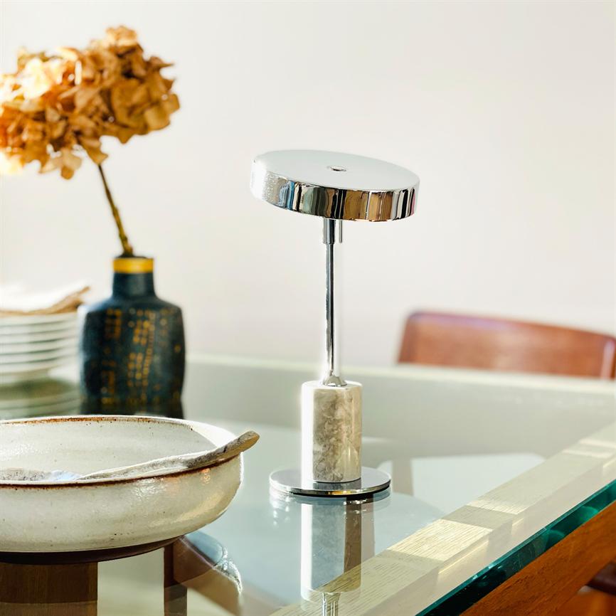 Eine moderne Tischlampe mit Chromscheibe und Marmorsockel spiegelt sich auf einem Glastisch wider, flankiert von einer Keramikschale und einer Vase mit Trockenblumen im Esszimmer.