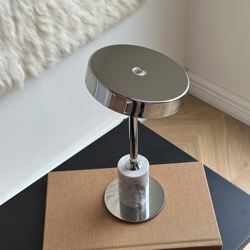 Ein runder Tischspiegel aus Chrom steht auf einem zylindrischen Marmorsockel, reflektiert das Licht um einen kleinen Metallring auf der Oberseite und liegt auf einem braunen Buch auf einem schwarzen Tisch in einer hellen Ecke mit Parkettboden.