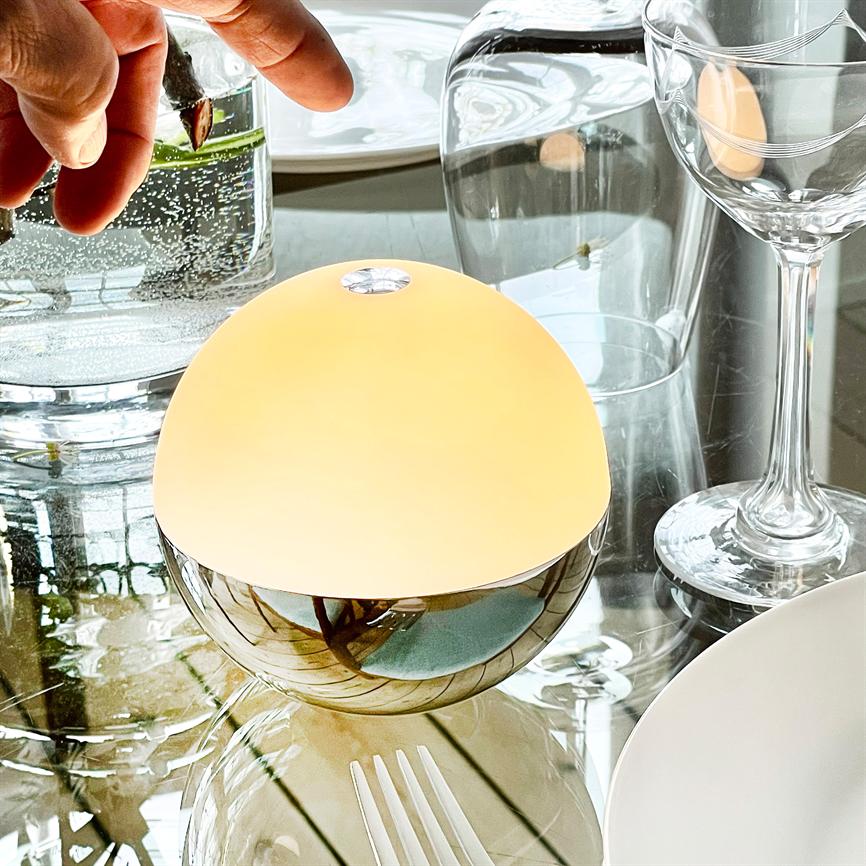 Eine runde Tischlampe leuchtet warm; es steht auf einem verspiegelten Glastisch, umgeben von Weingläsern, einem Teller, Gabeln und einem Glas mit einer Hand, die einen Pflanzenzweig hält.