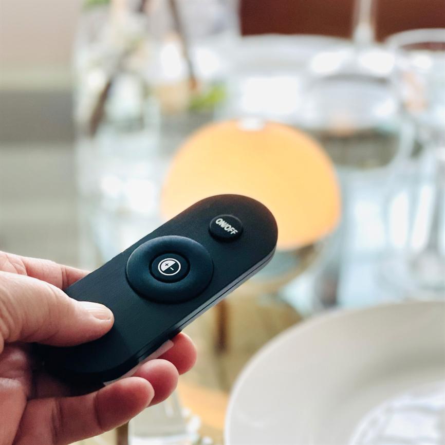Eine schwarze Fernbedienung wird in der Hand gehalten und auf eine warmorangefarbene Tischlampe gerichtet; Gläser und ein Teller liegen verschwommen auf einem gedeckten Tisch im Hintergrund.
 
 Text im Bild: „ON/OFF“ und „+“