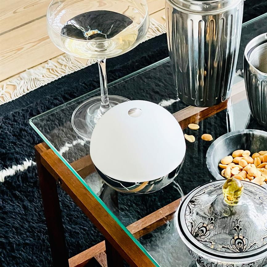 Der weiße, halbkugelförmige Diffusor steht auf einem Glastisch, umgeben von einem Weinglas, Metallbehältern und einer Schale mit Erdnüssen auf einem schwarzen, pelzähnlichen Teppich.