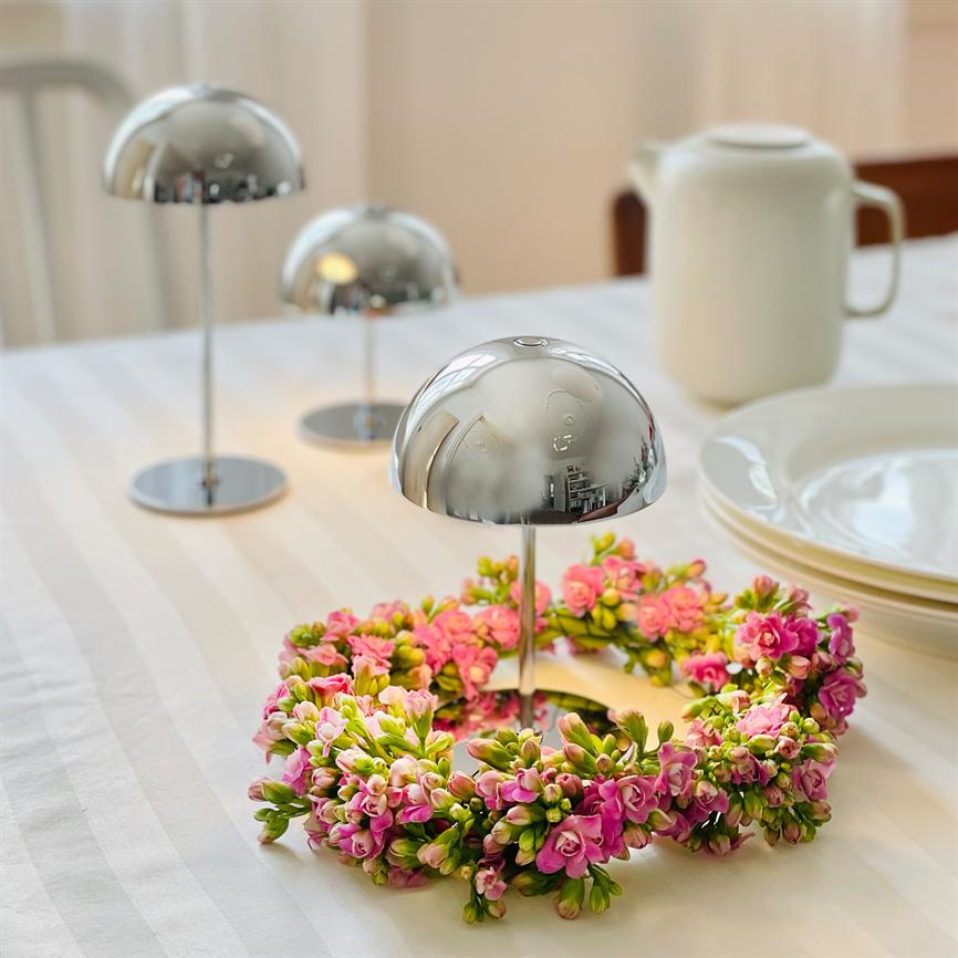 Silberfarbene, halbkugelförmige Ständer stehen auf einer weiß gestreiften Tischdecke; Eine Girlande aus kleinen rosa Blumen umgibt den vorderen Ständer, im Hintergrund sind gestapelte Porzellanteller und ein weißer Krug zu sehen.
