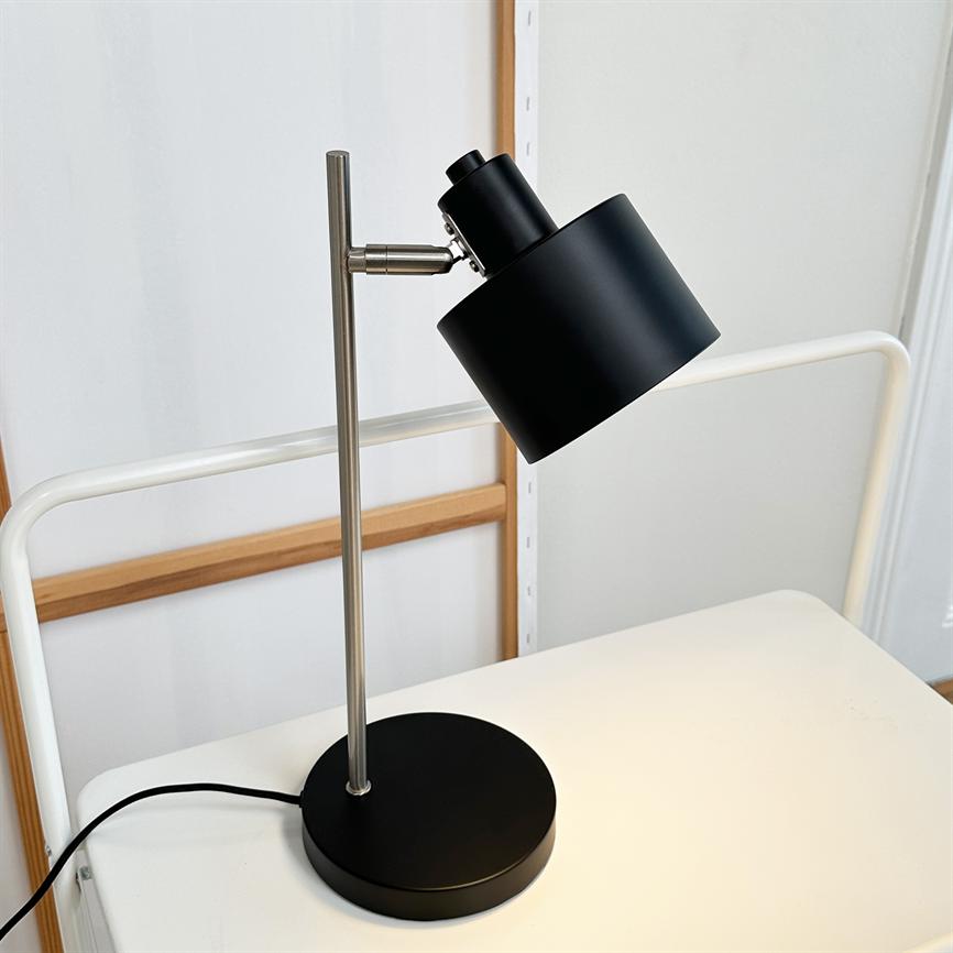 Schwarze Schreibtischlampe steht auf weißem Tisch; Der Schirm ist geneigt und wirft das Licht nach unten. Die Lampe hat einen silbernen Stab und einen runden schwarzen Sockel, vor einer weißen Wand mit Holzrahmen.