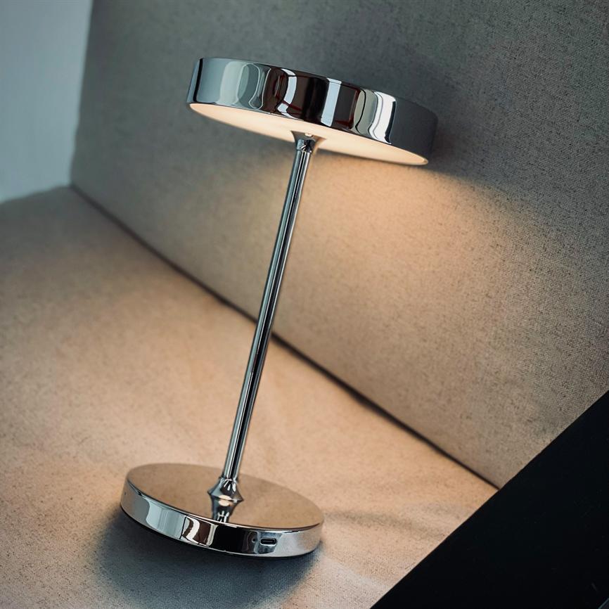 Eine verchromte Tischleuchte strahlt sanft nach unten, schräg geneigt; Die Lampe steht auf dem beige gepolsterten Sitz des Sofas an der Rückenlehne mit reflektierendem Metallsockel und rundem Schirm.