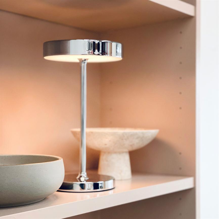 Eine verchromte Tischlampe wirft sanftes Licht auf ein hellrosa Regal neben einer runden grauen Schüssel und einer beigen Schüssel mit Fuß.