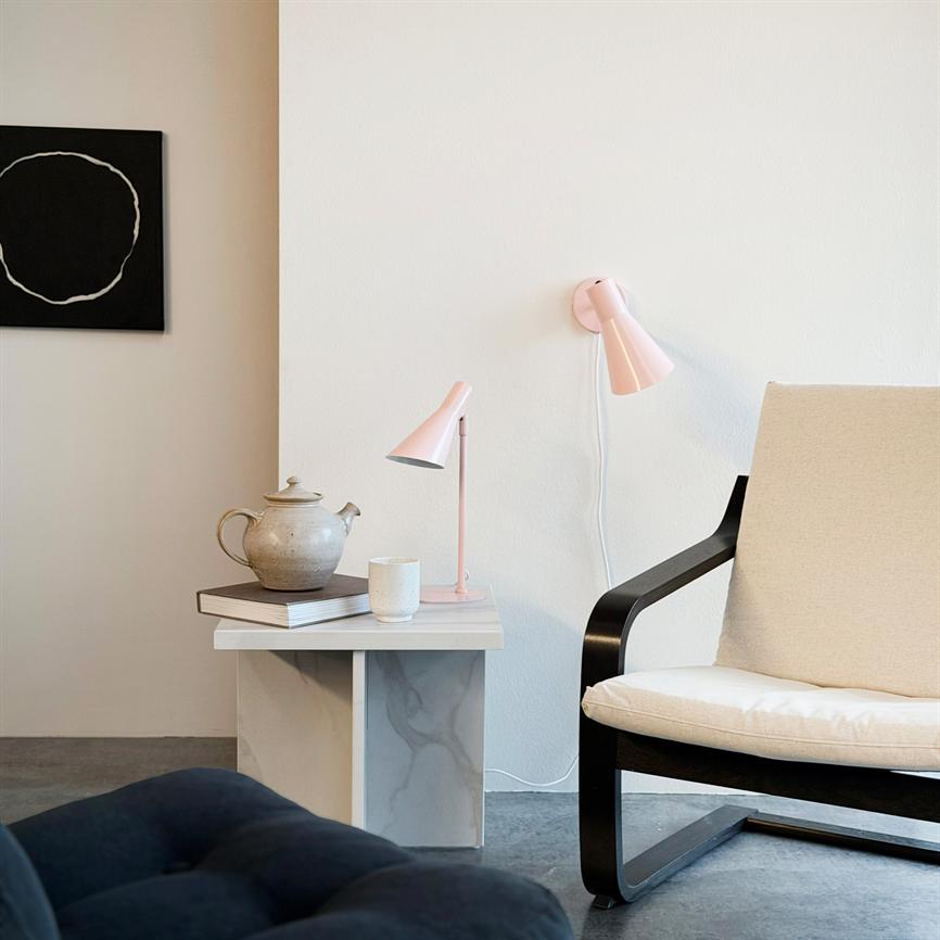 Ein beiger Sessel steht neben einem kleinen Marmortisch, auf dem eine Teekanne, eine Tasse und eine rosa Lampe stehen; Weiße Wand und ein schwarzes abstraktes Kreisgemälde in einem minimalistischen Raum.