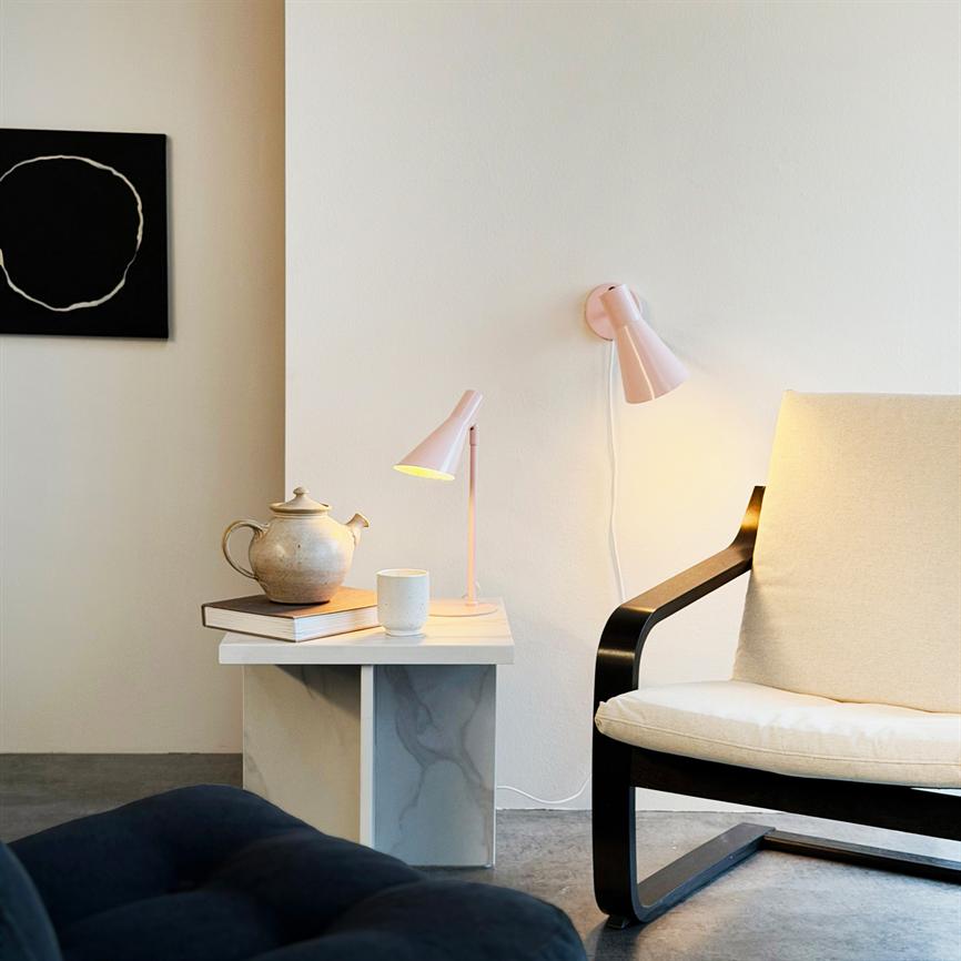 Sessel steht neben Beistelltisch; Auf dem Tisch stehen eine Teekanne, eine Tasse und ein Buch, eine Tischlampe leuchtet und eine Wandlampe verbreitet ein warmes Licht in einem hellen, minimalistischen Raum.