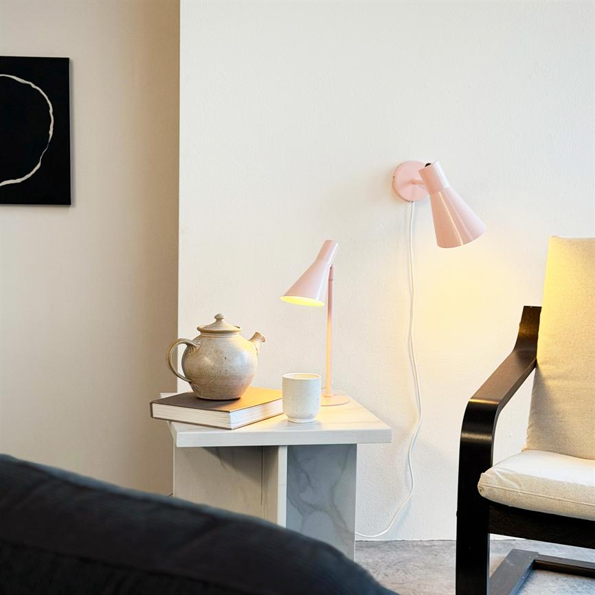 Auf einem Beistelltisch steht eine Teekanne aus Keramik, dazu eine Tasse und zwei rosafarbene Lampen, neben einer hellen Wand und in einer ruhigen Ecke ein Sessel mit schwarzen Armlehnen.