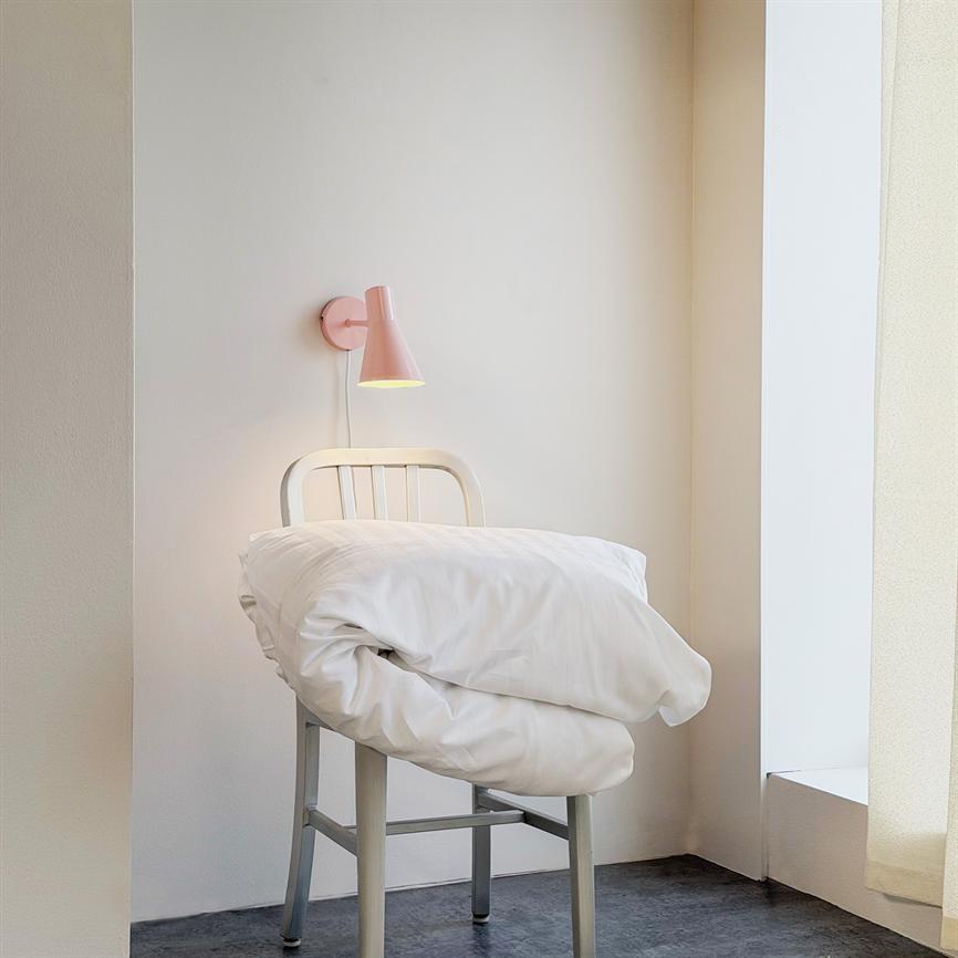 En sammenfoldet hvid dyne ligger på en grå metalstol, mens en pink væglampe lyser svagt i et lyst, minimalistisk hjørne ved en vindueskarm.