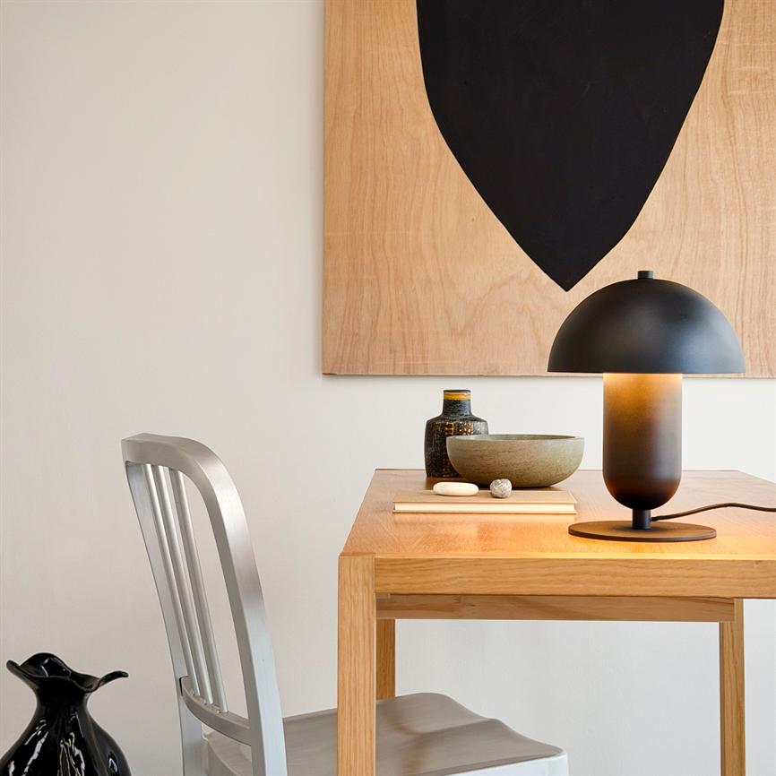 Eine mattschwarze Tischlampe wirft warmes Licht auf einen hellen Holztisch mit Keramikschale, Flasche und Buch; Am Tisch steht ein Metallstuhl und an der Wand hängt ein abstraktes Schwarzformatgemälde.